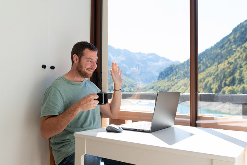a man in a virtual meeting with coworker holding a cup coffee while waving hello