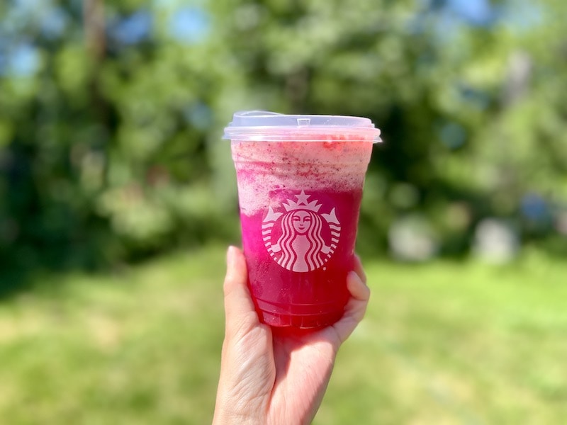 Starbucks Frozen Mango Dragonfruit Refresher drink held in front of summer yard